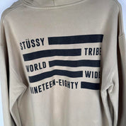 Stüssy oversized beige hoodie with Tribe spellout logo and relaxed fit.
